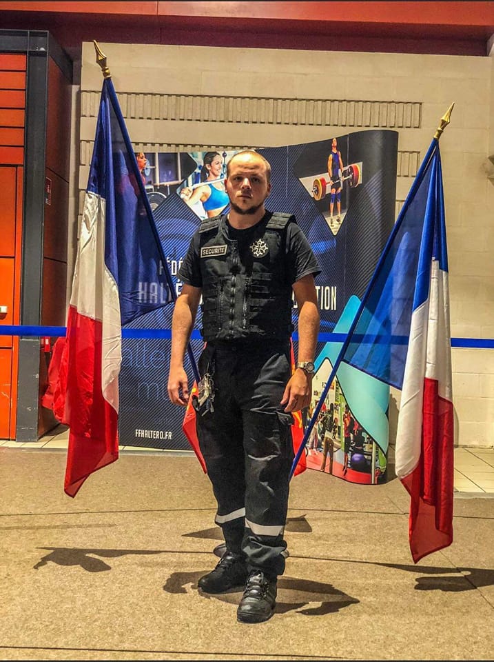 agent Pro Tech 12 en uniforme entre deux drapeaux à Villefranche-de-Rouergue devant panneau haltérophilie,